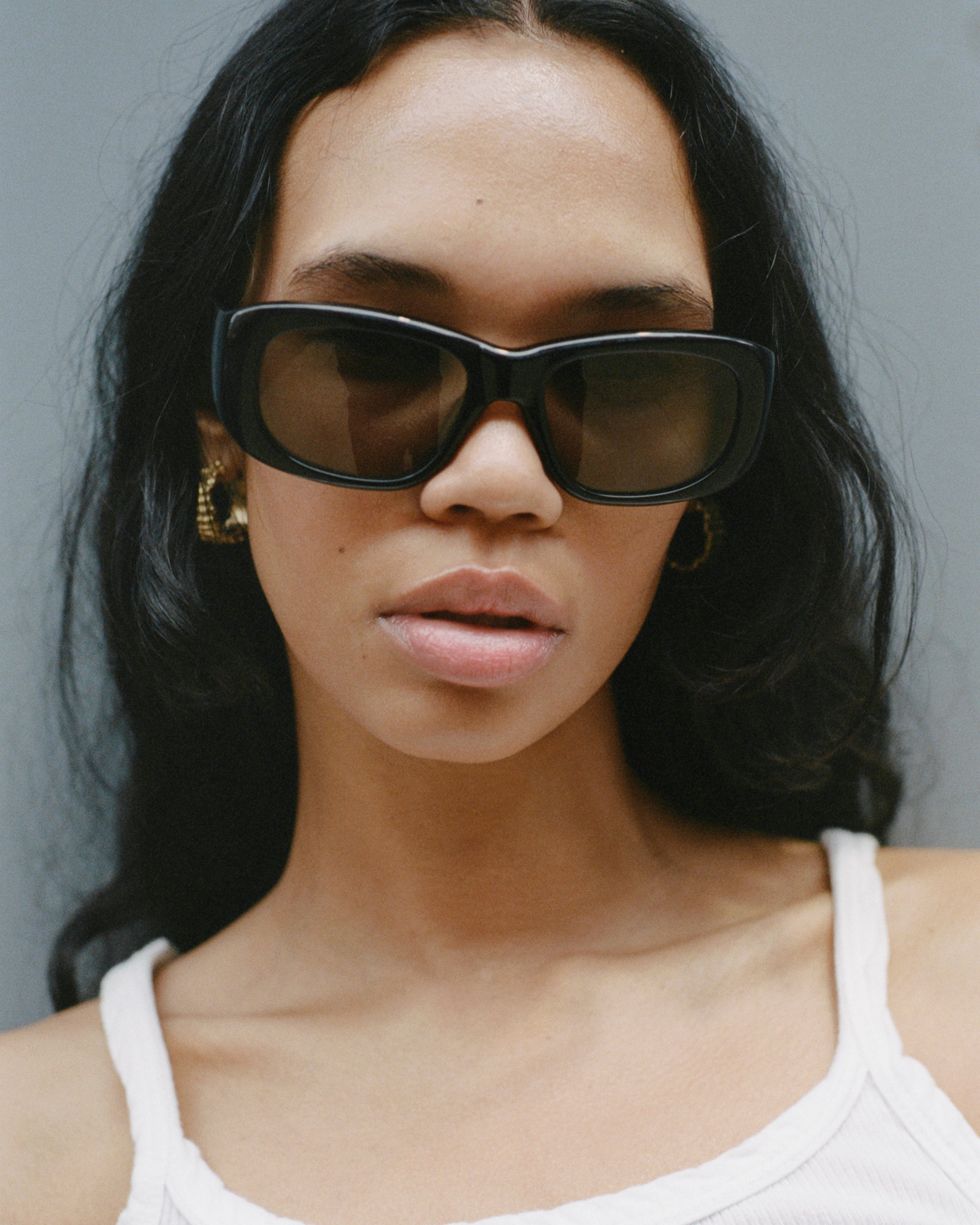 model wearing sunglasses, standing against a blue-grey wall