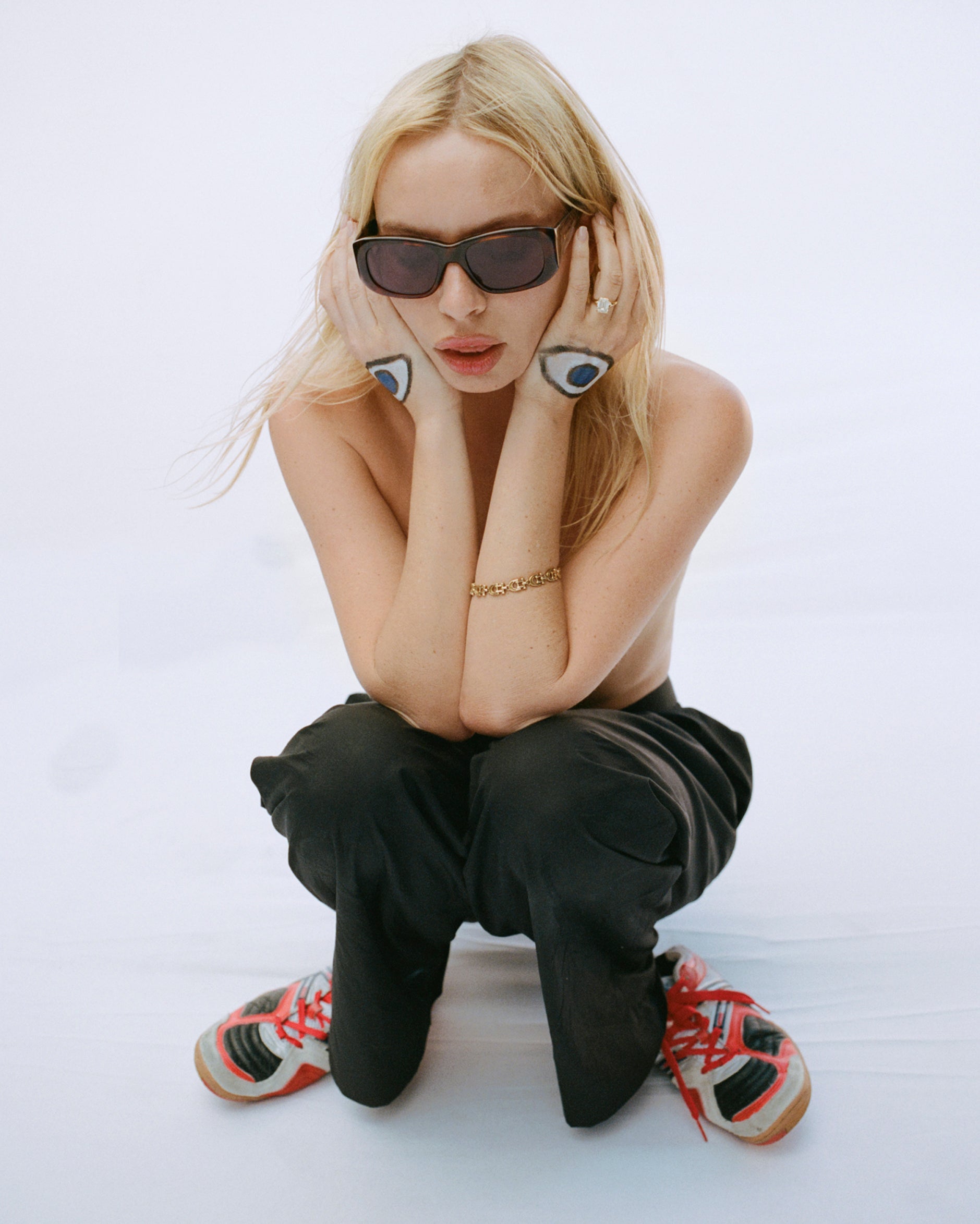 a model is crouched down and holding her face between her hands. there are eyes painted onto the backs of her hands.