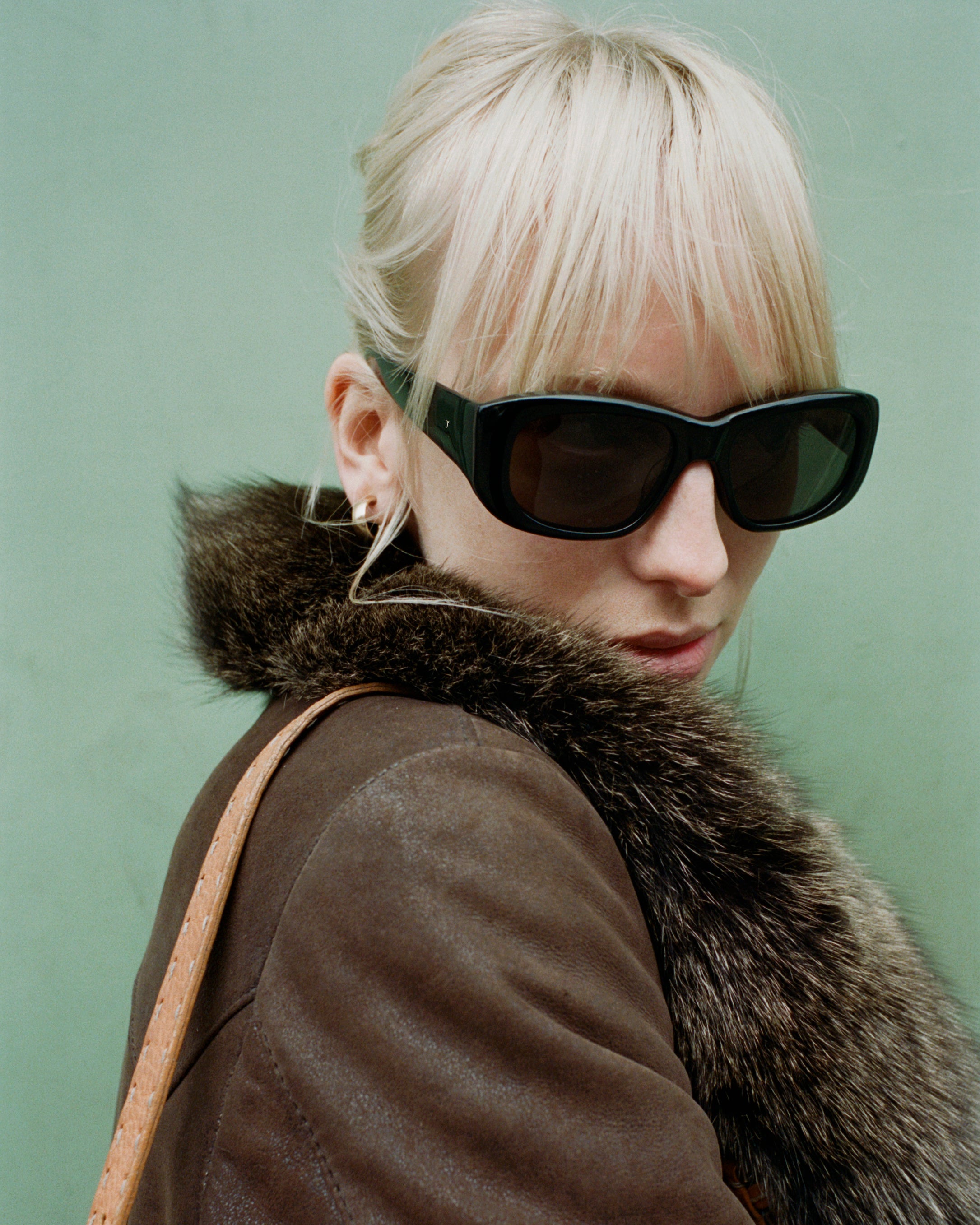 a model looking over her shoulder. she is standing in front of a sage green wall, wearing black sunglasses and a brown fur coat.