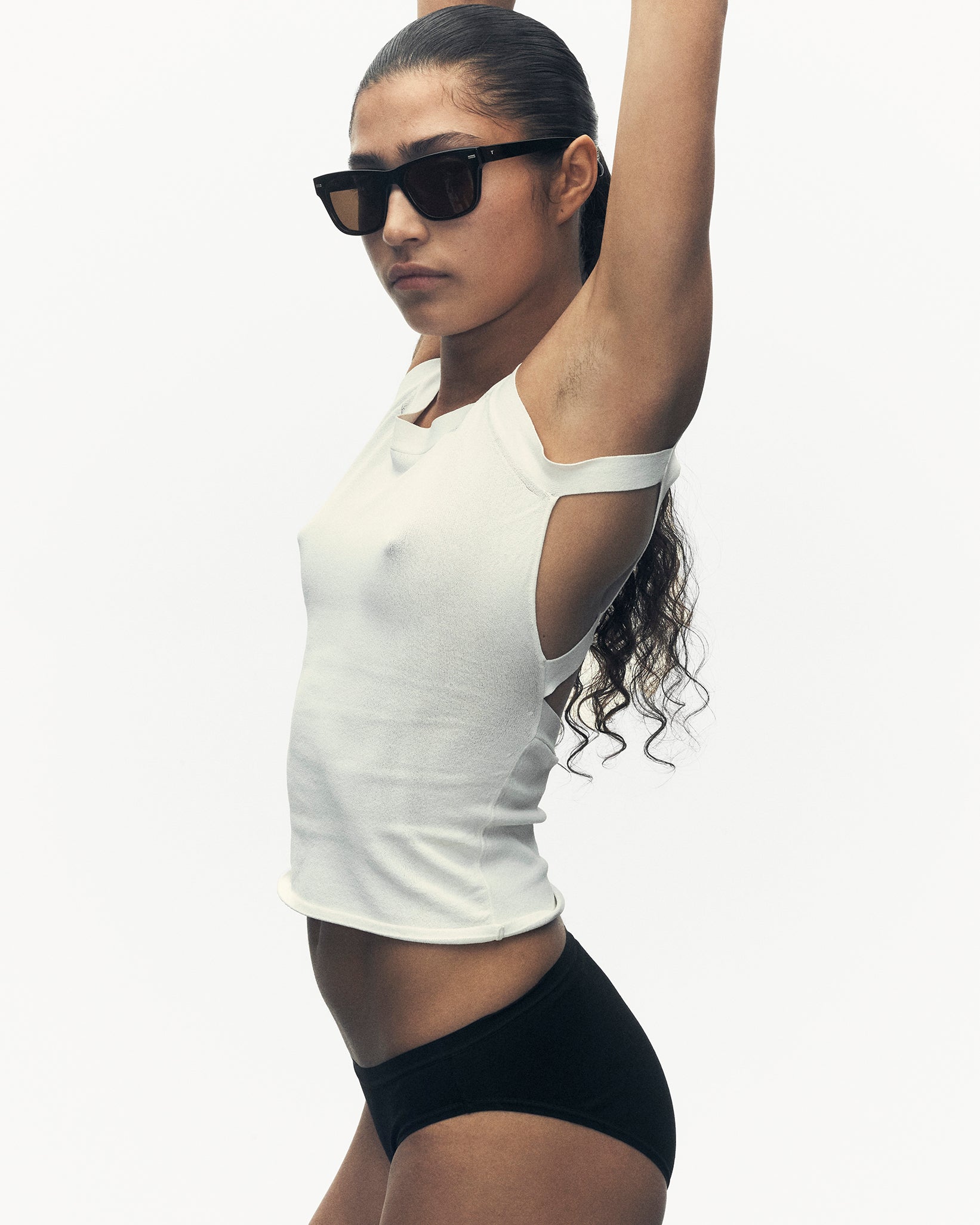 a model with her arms up. she is wearing sunglasses, a white tank top, and black panties.