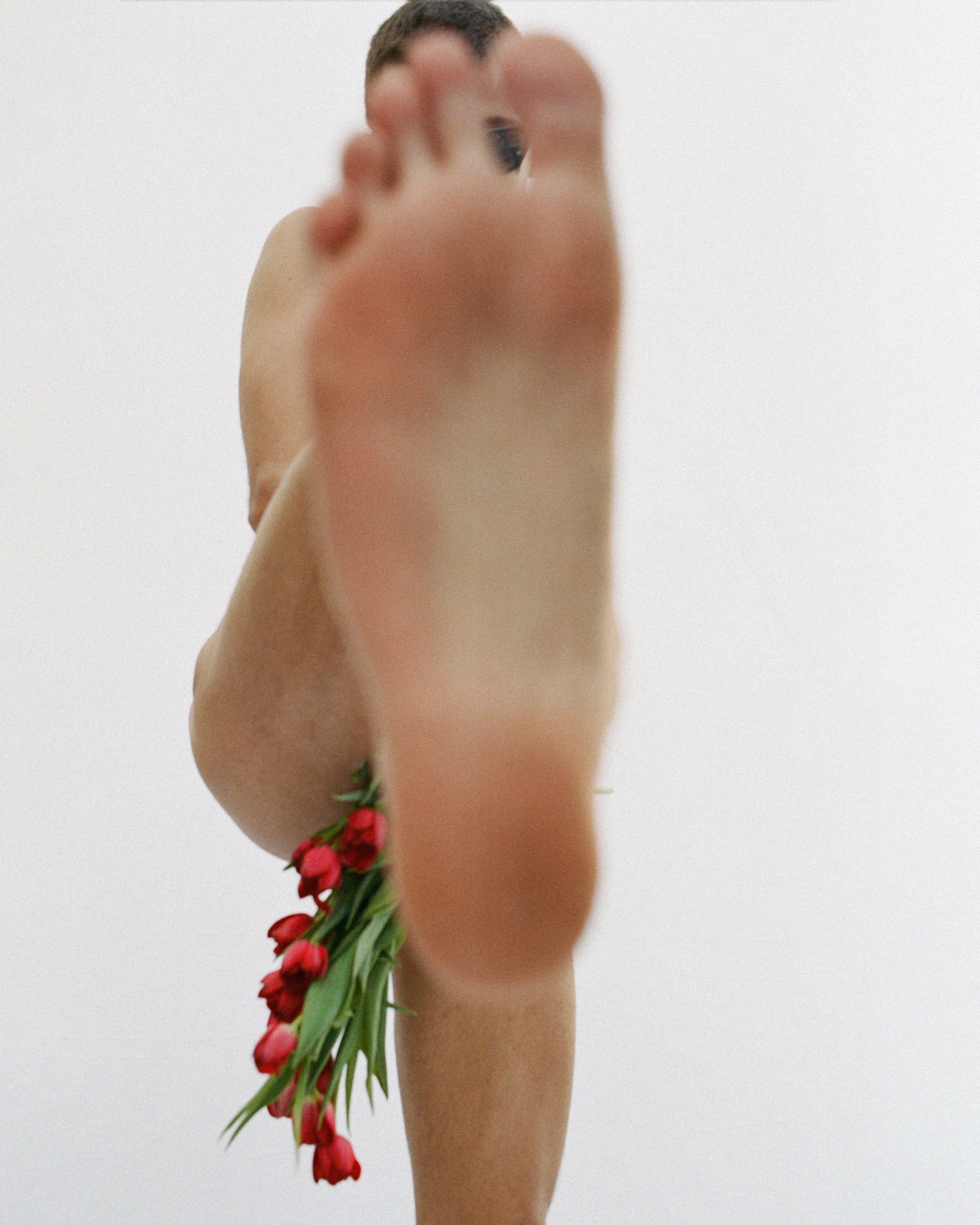 a nude person holding a bouquet of red flowers and kicking their leg up toward the camera. their bare foot obstructs most of the view of their body.
