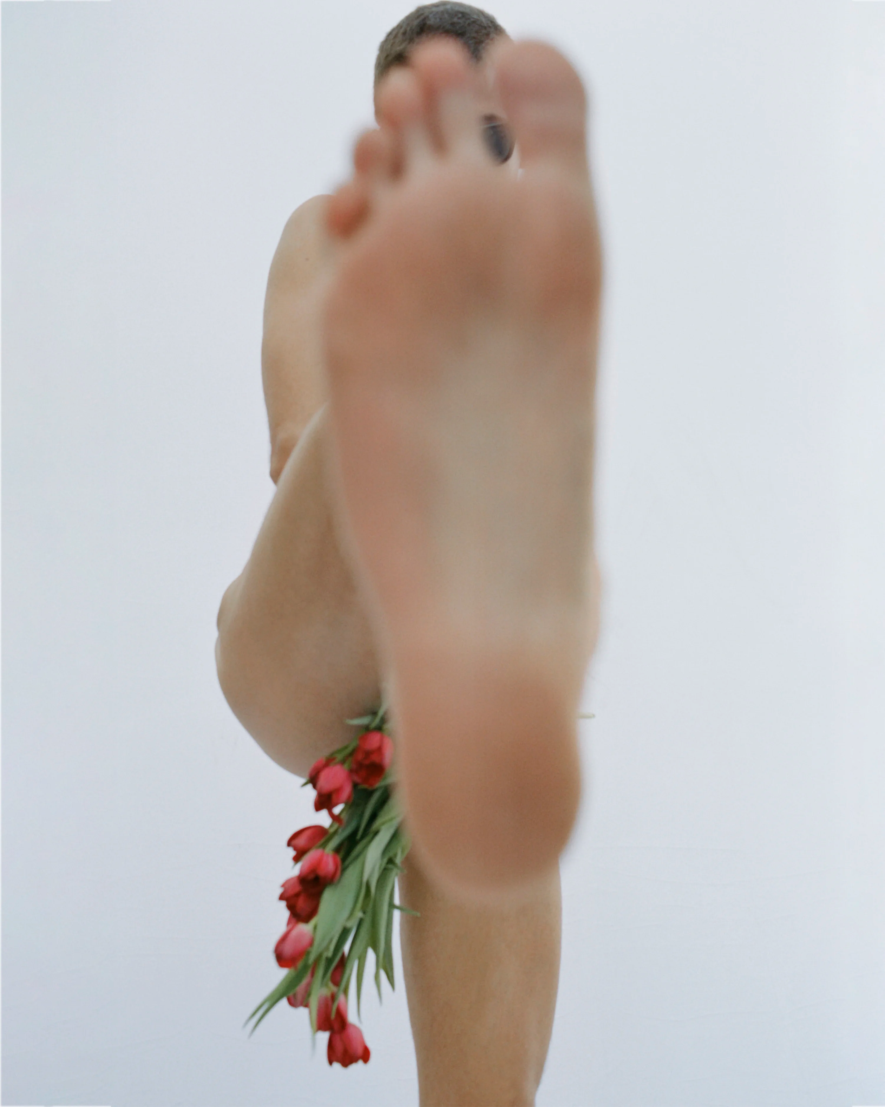 a nude person holding a bouquet of red flowers and kicking their leg up toward the camera. their bare foot obstructs most of the view of their body.